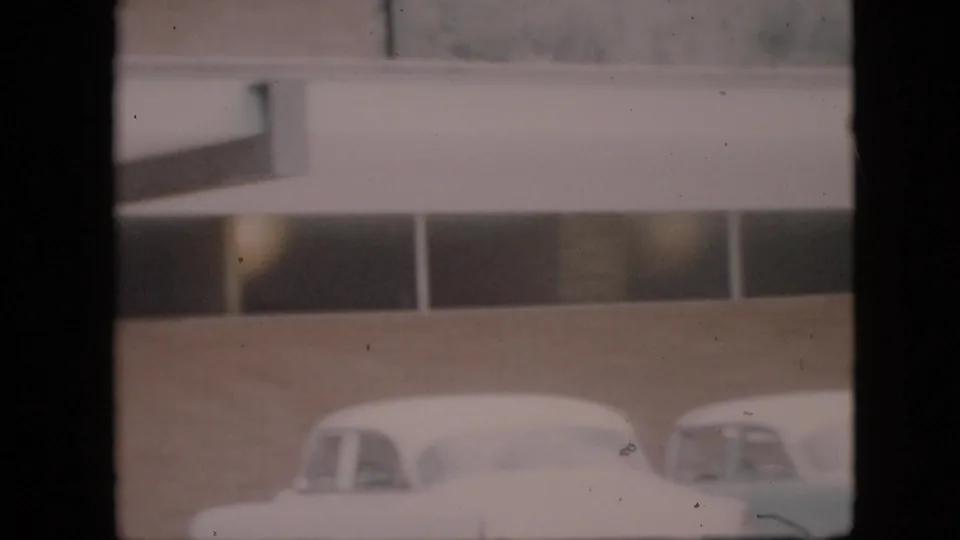 Two white cars appear partly framed in front of a brick building with a flat canopy and high rectangular windows in the 1958 Pennsylvania home-movie footage.