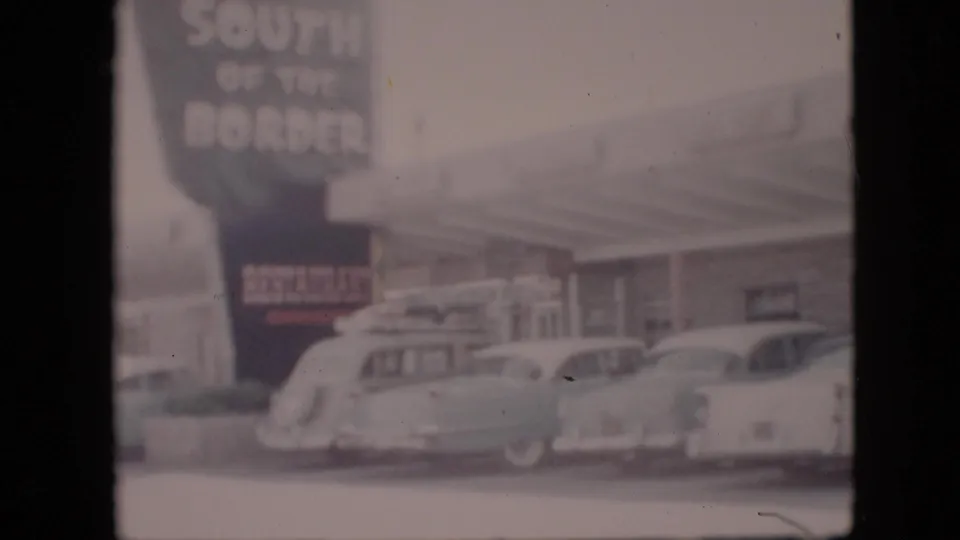 Soft 8mm view of a 1958 Pennsylvania roadside restaurant or gas station with a large South of the Border sign and several parked cars.