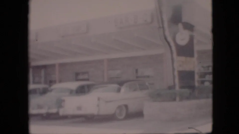 Faded 8mm frame from the 1958 Pennsylvania roadside stop, with classic cars, a covered storefront, and partial restaurant or service signage visible.