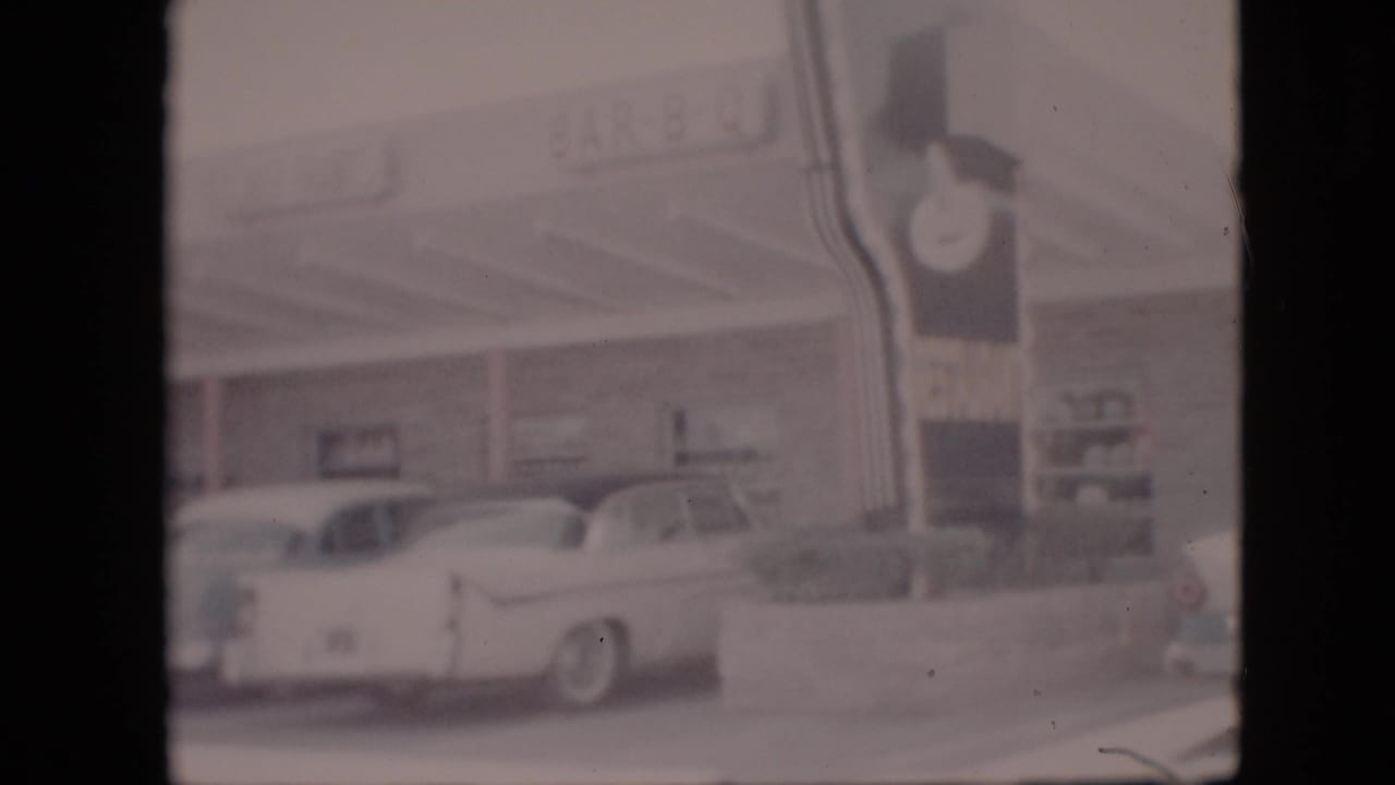 1958 Pennsylvania home-movie frame showing a roadside BAR-B-Q restaurant exterior, a white 1950s sedan, curb plantings, and a tall sign with a clock.