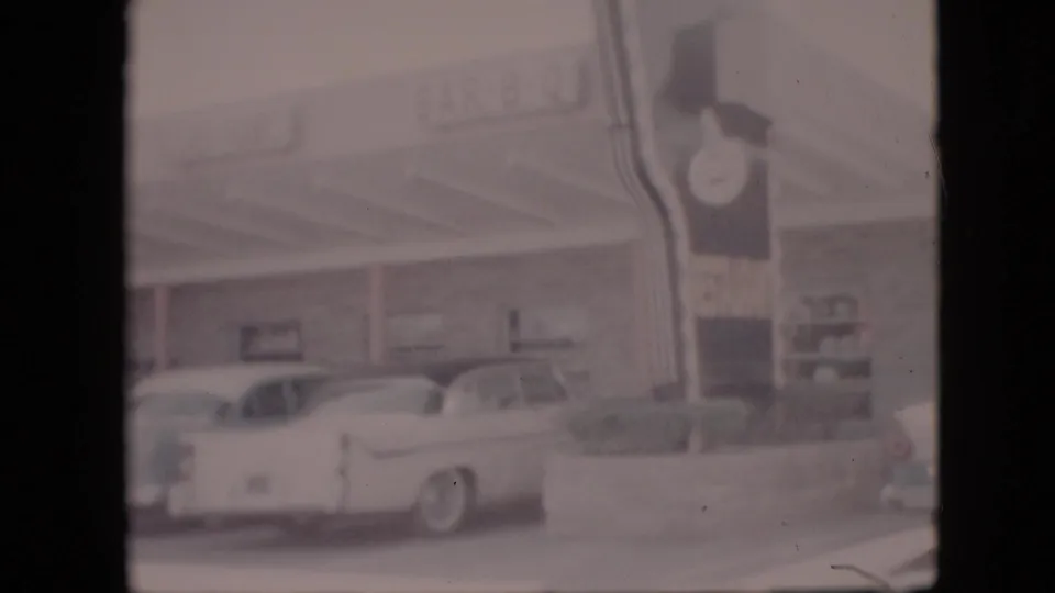1958 Pennsylvania home-movie frame showing a roadside BAR-B-Q restaurant exterior, a white 1950s sedan, curb plantings, and a tall sign with a clock.