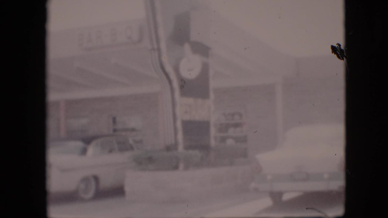 Soft 8mm frame of a Pennsylvania roadside BAR-B-Q restaurant in 1958, with the clock sign, parked white sedan, and another vehicle partly visible at the right edge.