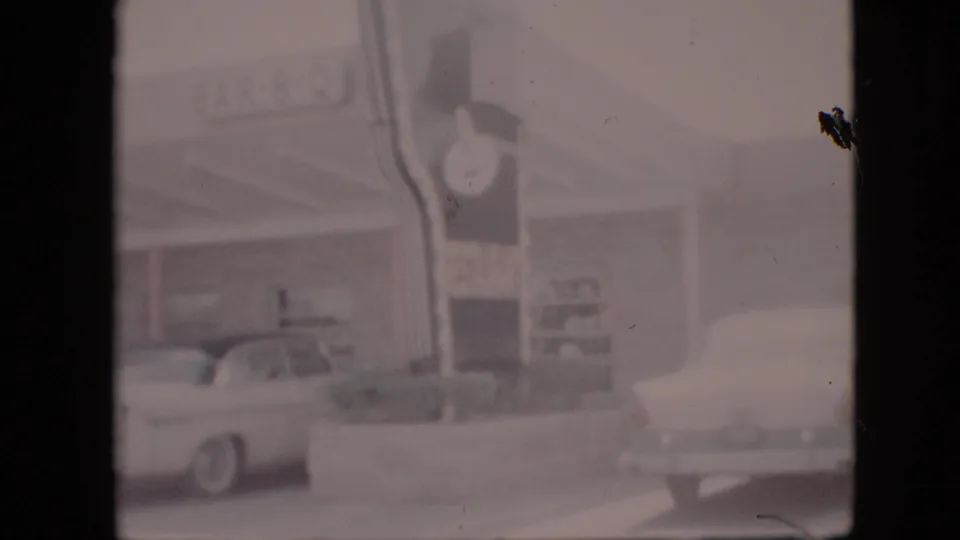 Soft 8mm frame of a Pennsylvania roadside BAR-B-Q restaurant in 1958, with the clock sign, parked white sedan, and another vehicle partly visible at the right edge.