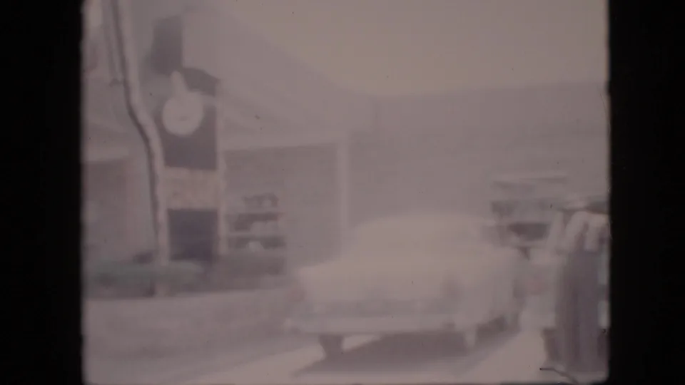 Home-movie frame from the 1958 Pennsylvania clip showing a light-colored 1950s car from the rear, the BAR-B-Q clock sign, restaurant frontage, and roadside curb area.