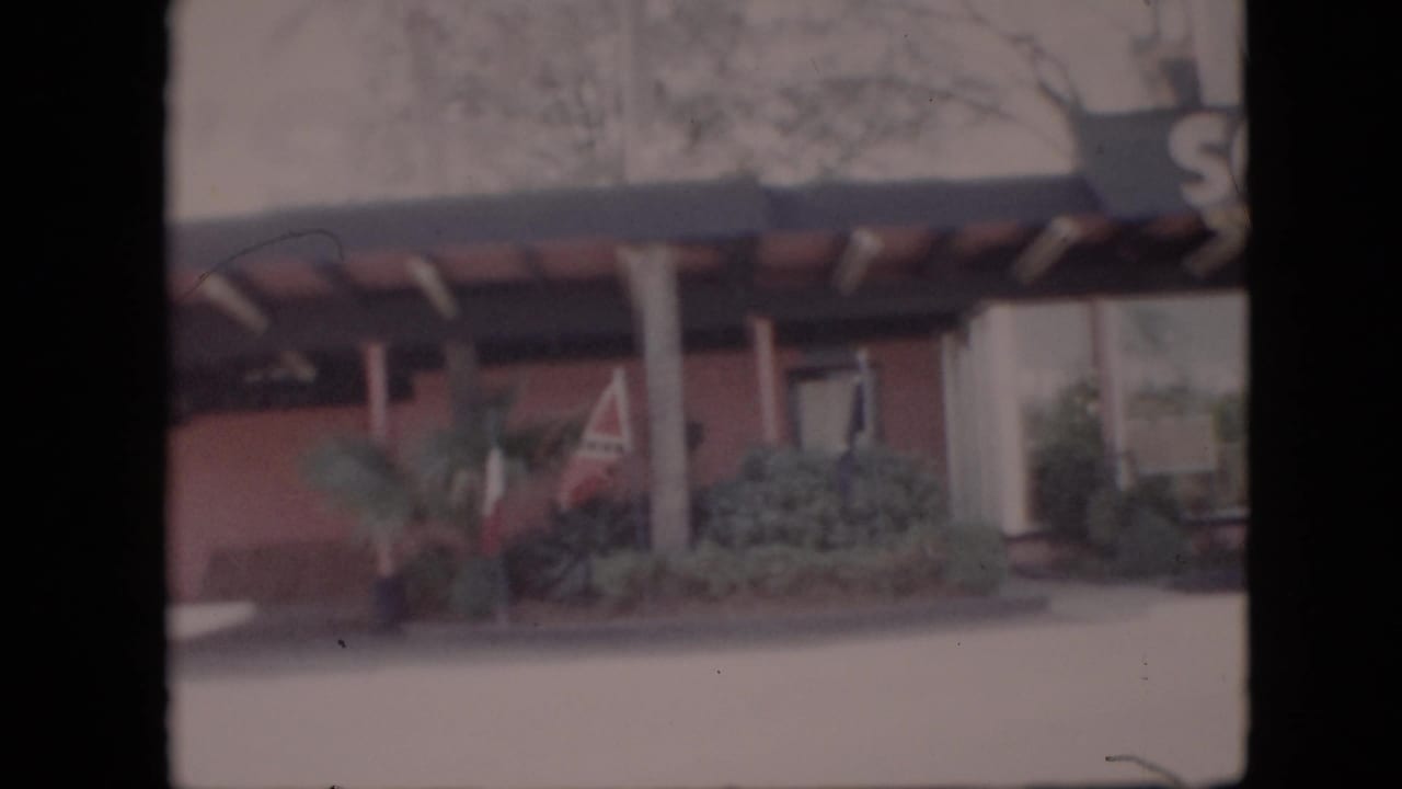 1958 Pennsylvania 8mm frame showing a mid-century building exterior with a covered entrance, support columns, shrubbery, and a small flag near the facade.