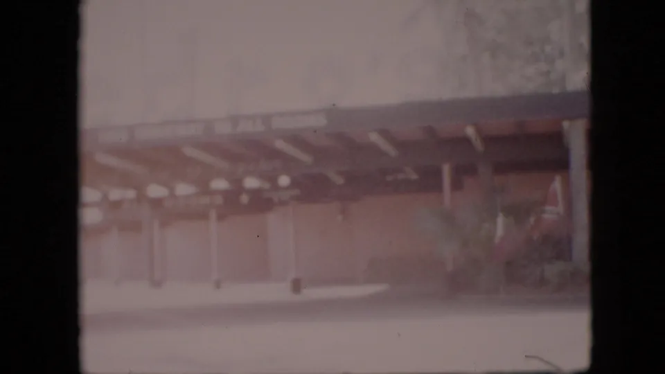 1958 Pennsylvania 8mm frame showing the building's covered entrance from across a drive, with columns, shrubbery, and a small flag partly visible.