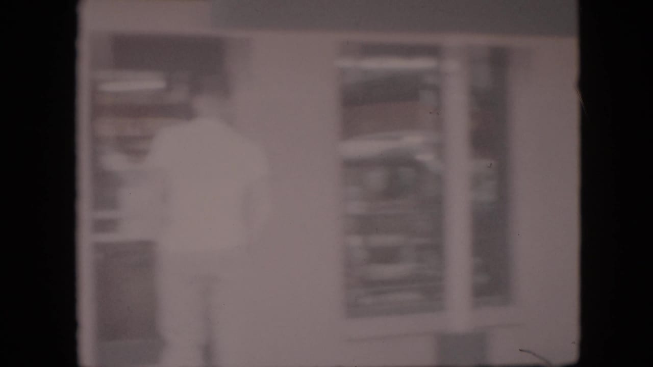 In this 1958 Pennsylvania 8mm home-movie frame, a light-clothed man appears near a doorway or interior opening, seen through heavy blur and overexposure.