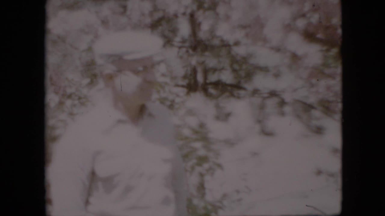 In 1958 Pennsylvania home-movie footage, a middle-aged man in a light shirt and cap appears partly hidden among pale flowering branches.