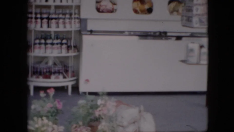Inside the Pennsylvania country store, a white refrigerated case stands behind shelves of bottled and packaged goods, with floral arrangements low in the foreground.