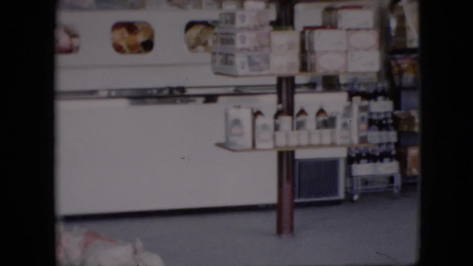 A mid-century store interior view shows a white refrigerated display case, stacked packaged goods, bottles, and shelves arranged along the wall.