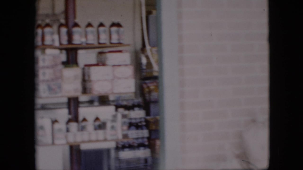 1958 Pennsylvania 8mm frame showing grocery shelves with bottles and canned goods near a brick wall and white sacks of bulk items.