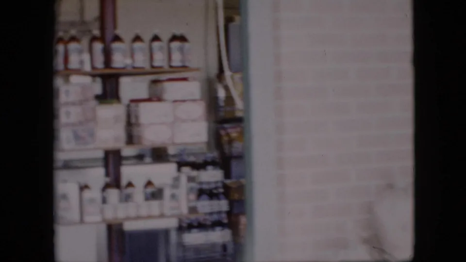 1958 Pennsylvania 8mm frame showing grocery shelves with bottles and canned goods near a brick wall and white sacks of bulk items.