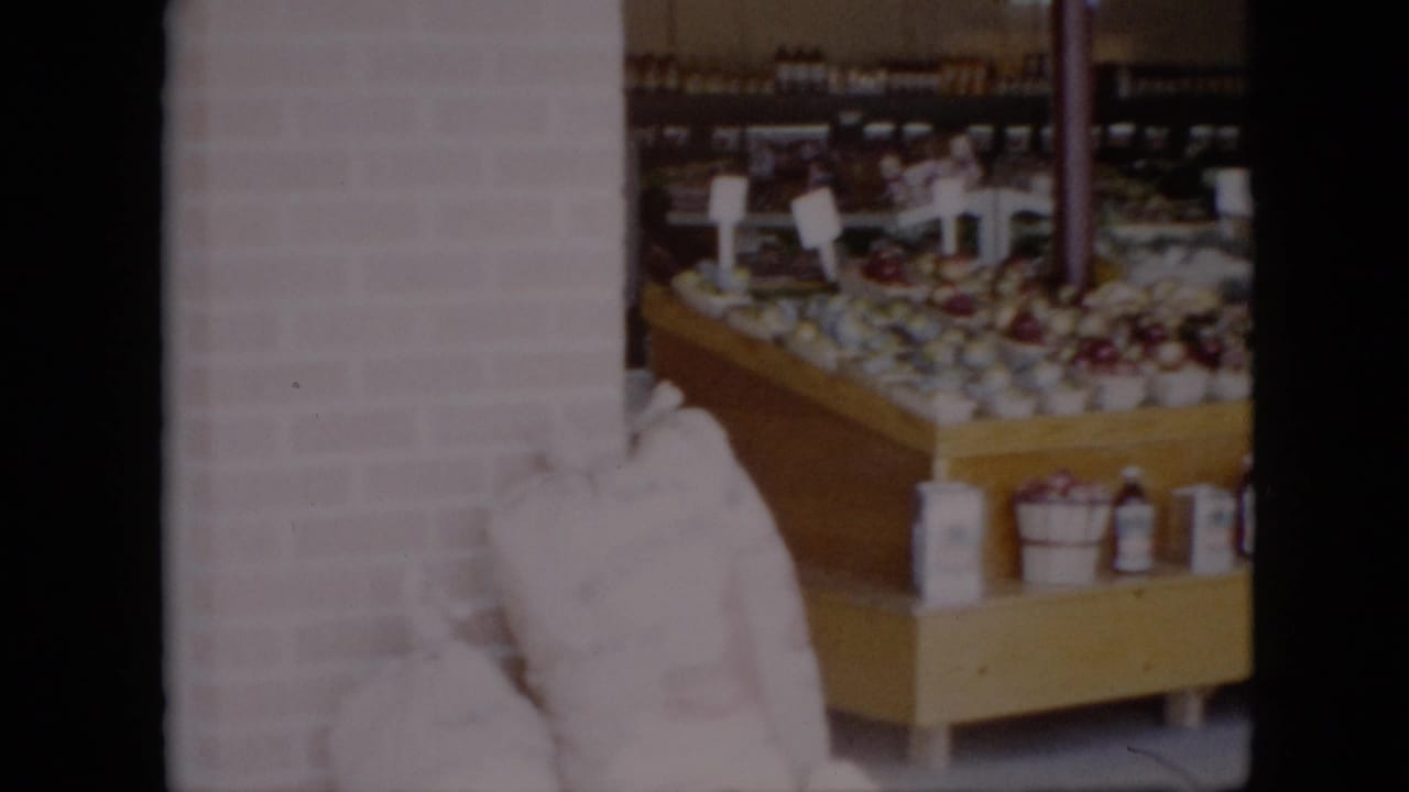 Produce display with baskets of fruit, bottles, cans, and white sacks near a brick wall.