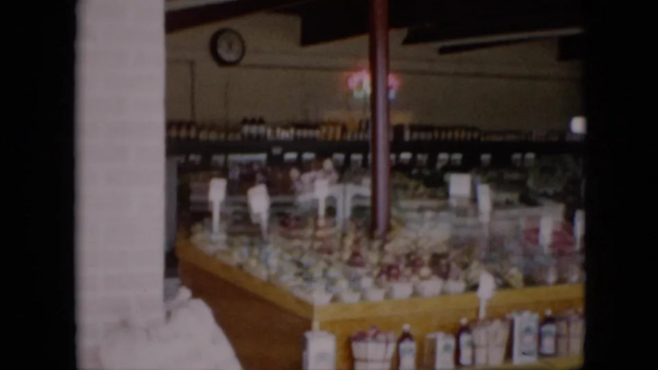 Dim 1958 Pennsylvania 8mm view of a grocery produce display with fruit baskets, bottles, white sacks, shelves, and a wall clock in the background.