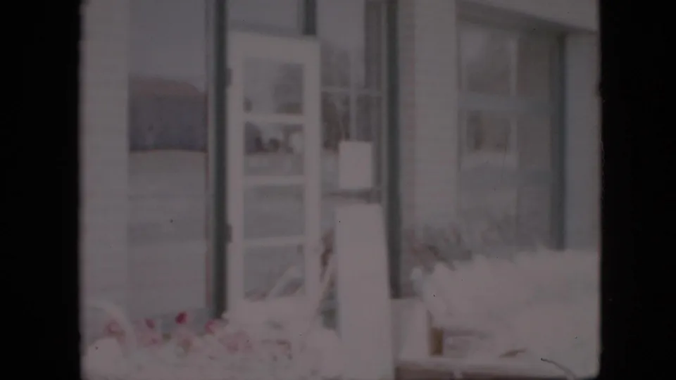 1958 Pennsylvania home-movie frame showing a grocery entrance area with glass panels, an open door, window framing, and flowers near the storefront.