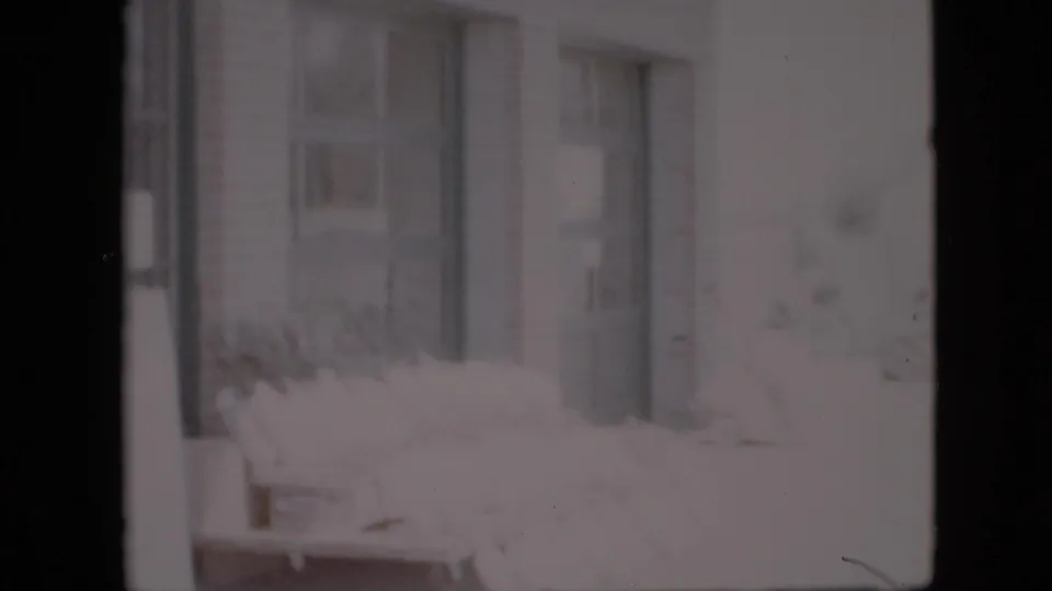 Soft, bright 8mm frame of the Pennsylvania grocery storefront with window panels, vertical framing, and washed-out flowers near the lower edge.