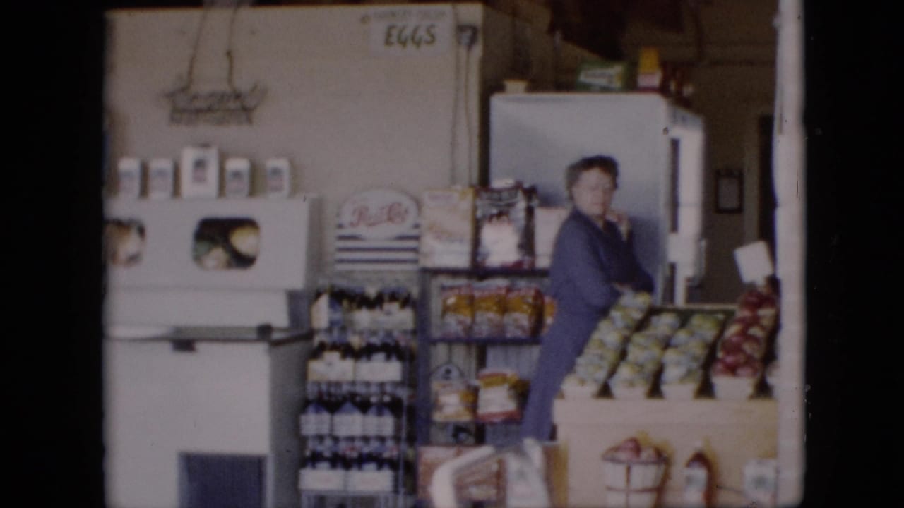 Woman in a blue dress near produce bins, bottles, shelves, and a white refrigerator.