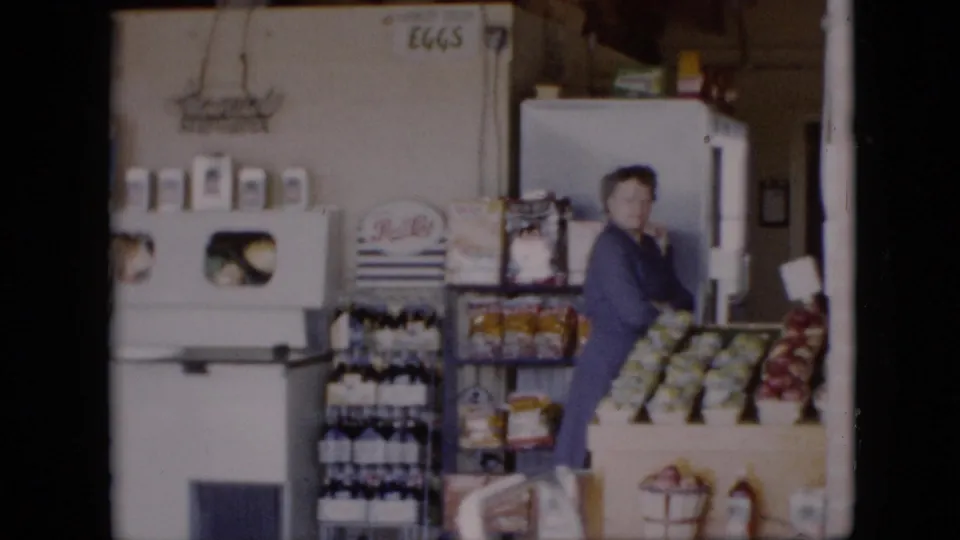 1958 Pennsylvania grocery-store frame showing a woman in a blue dress beside produce displays, bottles, shelves, and a white refrigerator or case.