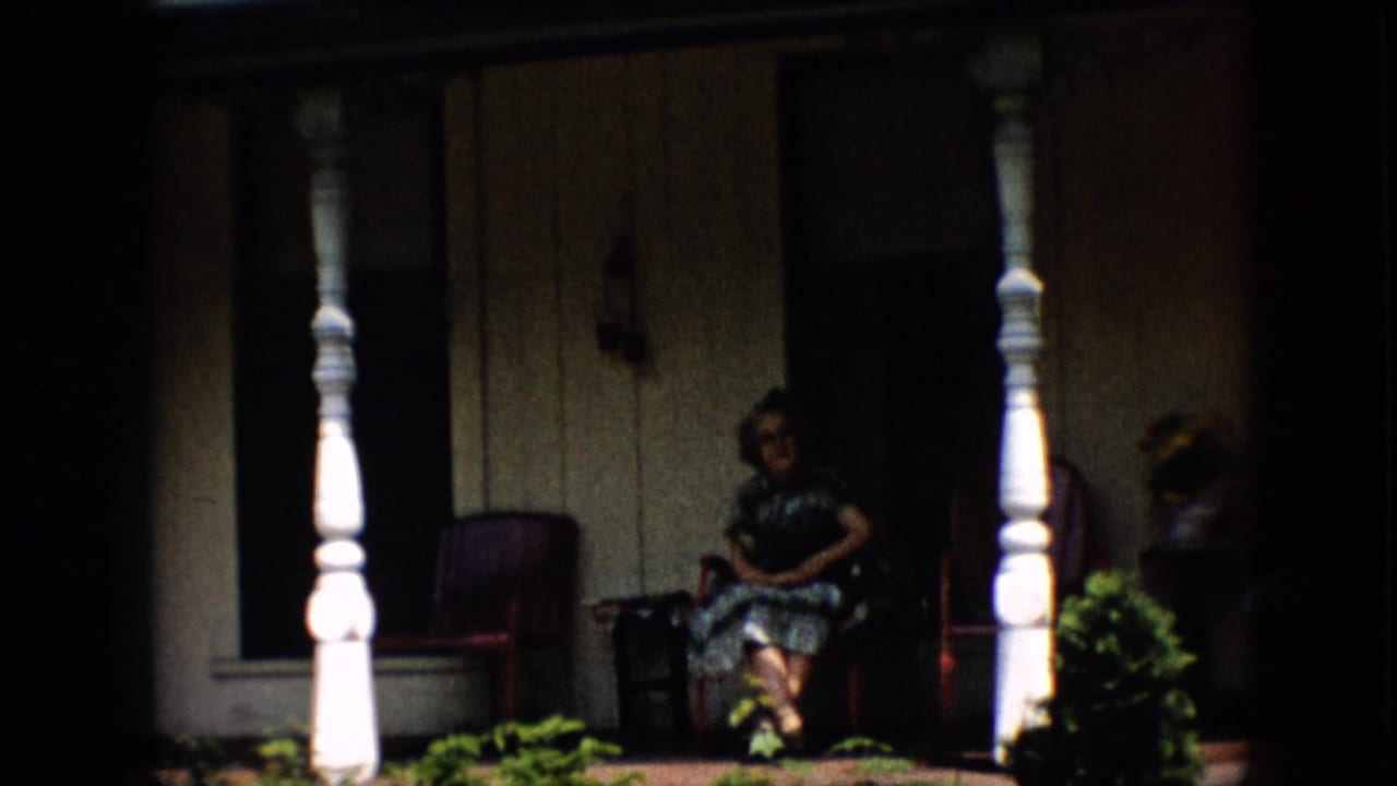 1958 Pennsylvania home-movie frame showing a woman with glasses seated on a shaded residential porch framed by white turned columns, red chairs, siding, and plants.