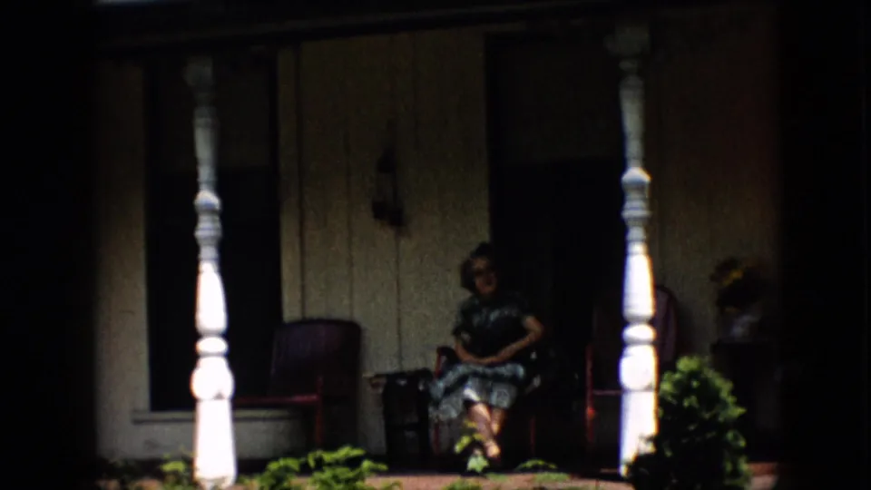 1958 Pennsylvania home-movie frame showing a woman with glasses seated on a shaded residential porch framed by white turned columns, red chairs, siding, and plants.