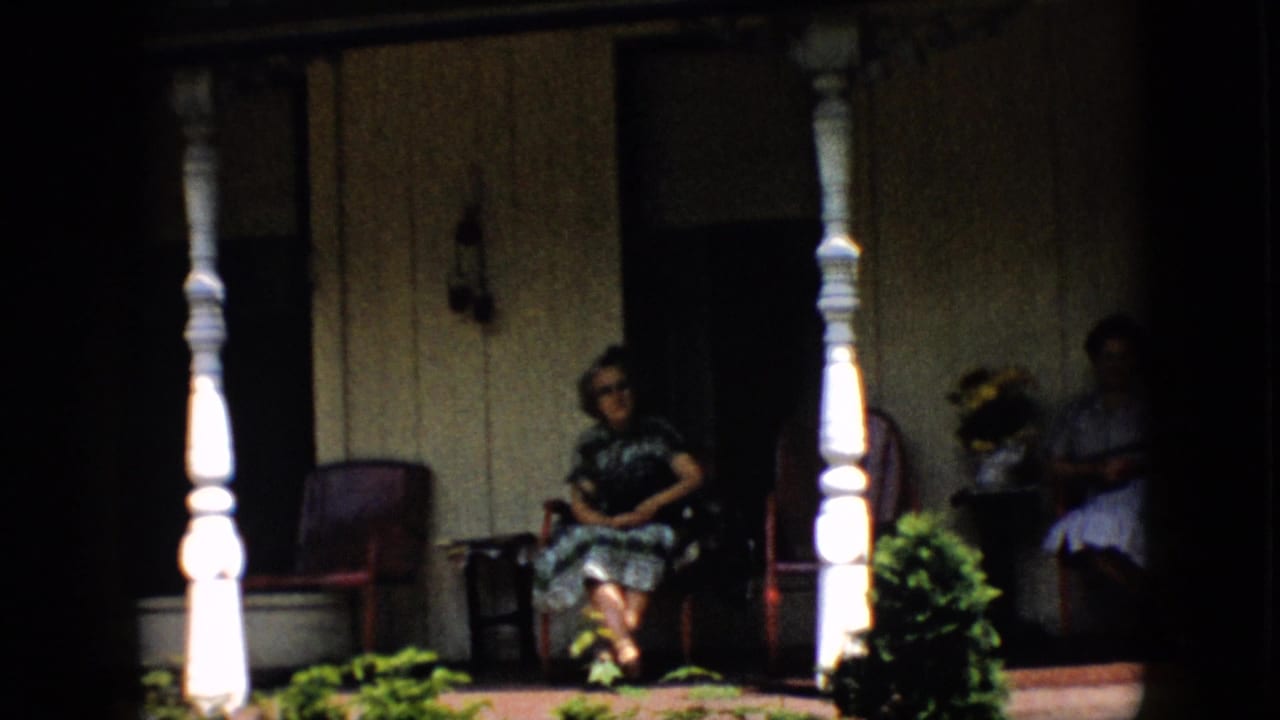 Two women seated apart on a shaded porch with white columns, red chairs, and plants.
