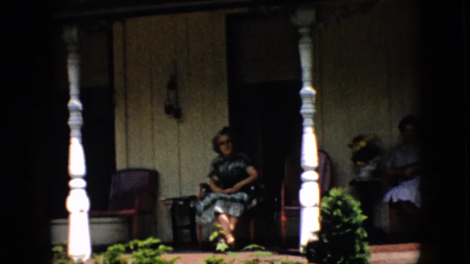 1958 Pennsylvania porch scene with one woman in a dark patterned dress seated near the center and another woman in a light dress seated near the doorway.