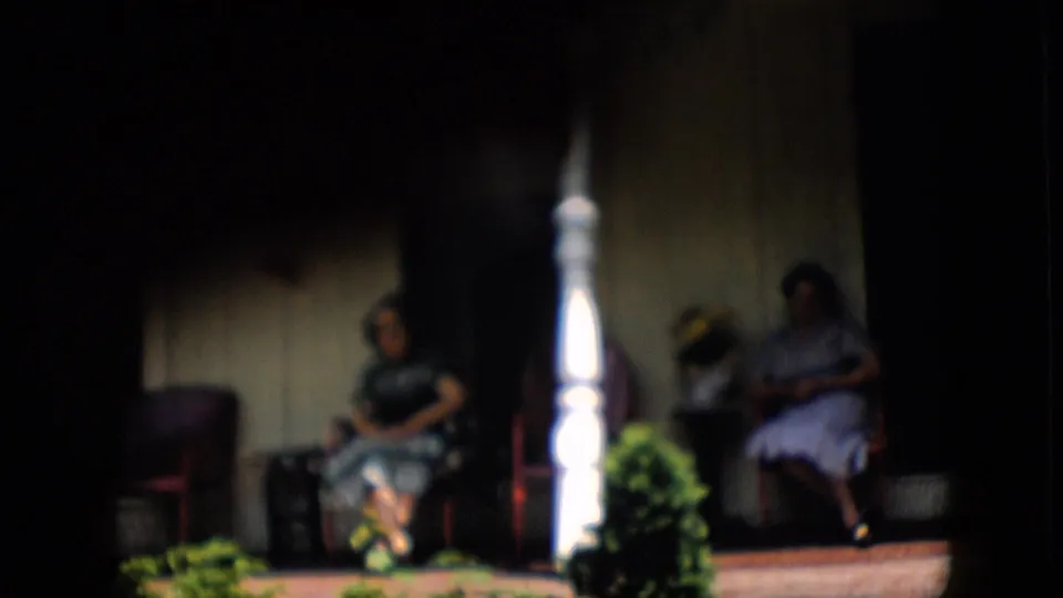Soft-focused 1958 Pennsylvania home-movie frame of two women seated on a shaded porch, partially obscured by deep shadow and a bright white column.