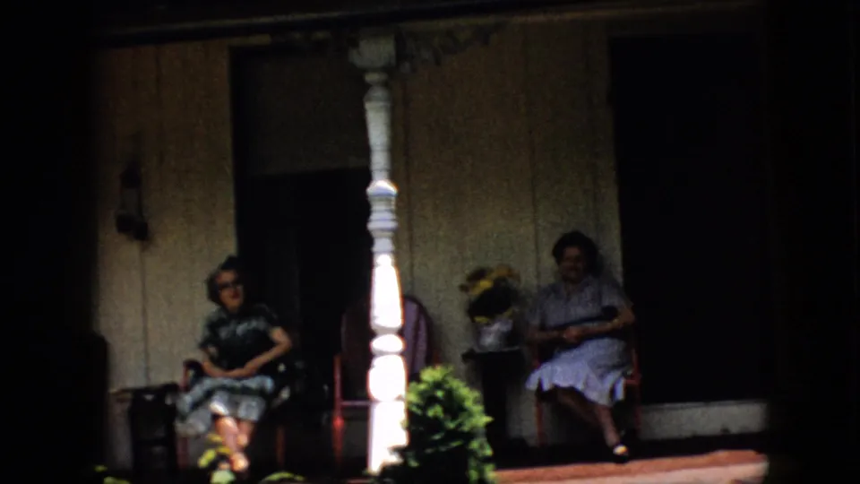 1958 Pennsylvania frame showing two women seated in shade on a residential porch with white columns, red chairs, potted flowers, and wood siding.