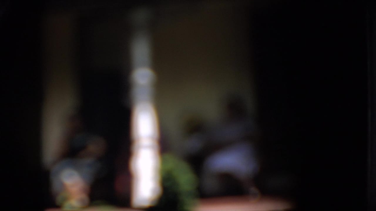A soft 1958 Pennsylvania 8mm frame shows two women seated on a front porch, partially obscured by a bright white spindle column and dark porch shadows.