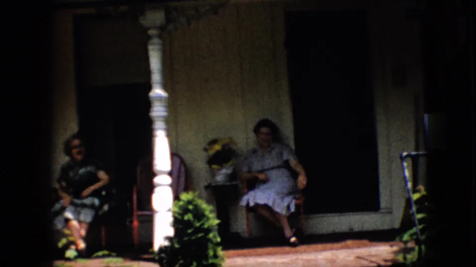 A clearer 1958 Pennsylvania home-movie frame shows two women in dresses seated in wooden chairs on a front porch with a white spindle column, potted plants, and a dark doorway or window behind them.