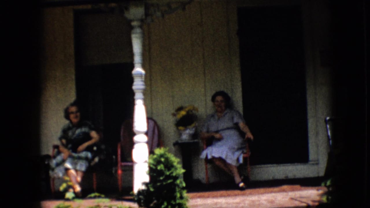 In this 1958 Pennsylvania home-movie frame, two adult women sit on a covered front porch, one in a dark patterned dress and glasses and the other in a light dress, with plants and white columns nearby.
