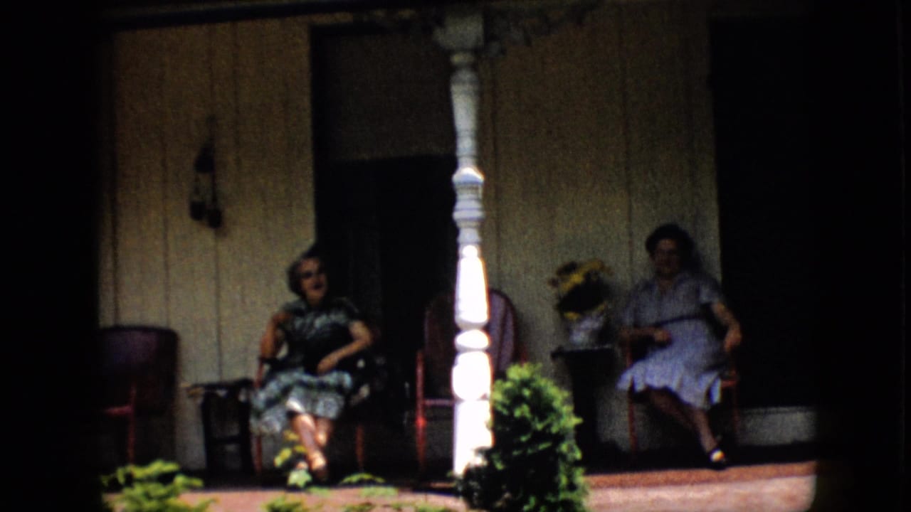 Two women remain seated on the shaded porch in Pennsylvania, framed by a white column, red-toned chairs, potted flowers, green shrubs, and dark window openings.