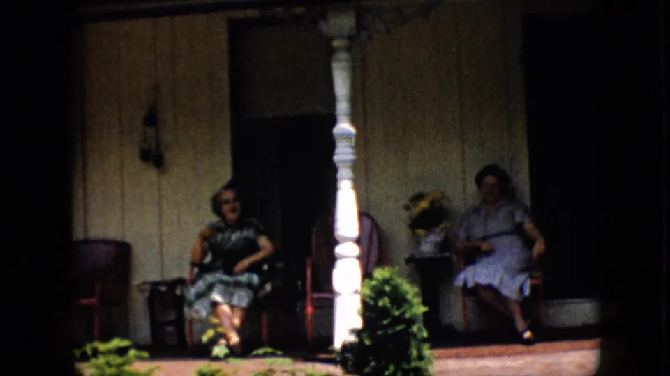 Two women remain seated on the shaded porch in Pennsylvania, framed by a white column, red-toned chairs, potted flowers, green shrubs, and dark window openings.