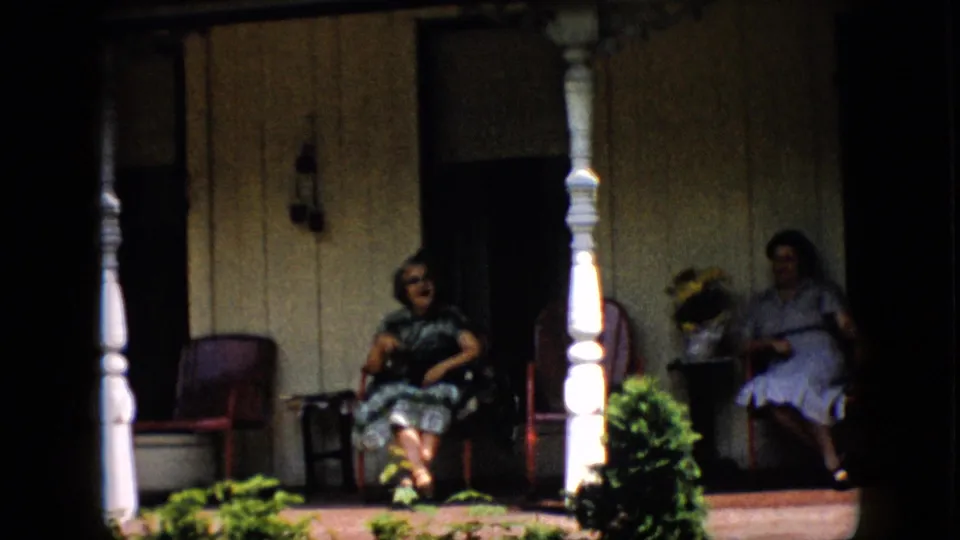 The 1958 home-movie frame shows one woman in a dark patterned dress and glasses near the center of the porch while another woman sits farther right in shadow, with white columns and plants in the foreground.