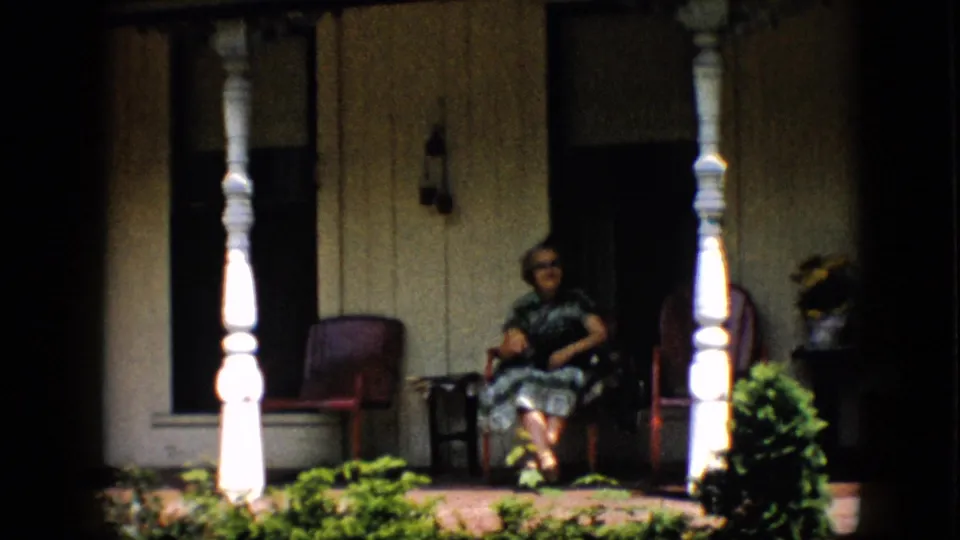 At the end of the clip, a woman in a dark patterned dress sits on the covered porch near a white column, while the second woman is partly visible at the right edge near a potted plant and shaded doorway.