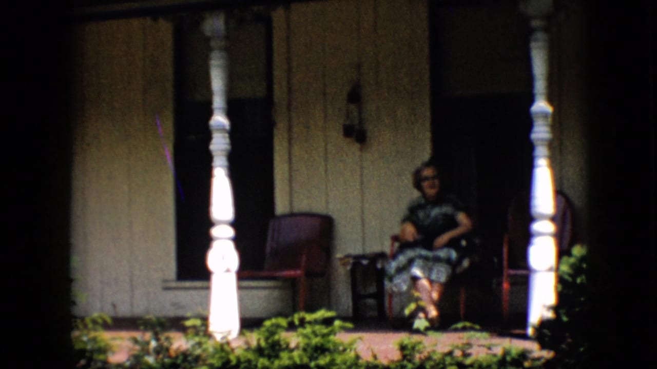 In 1958 Pennsylvania home-movie footage, a woman in glasses and a patterned dress sits on a front porch between white posts and red chairs.