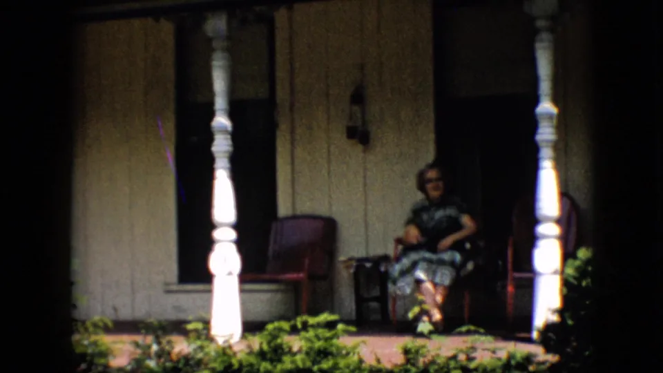 In 1958 Pennsylvania home-movie footage, a woman in glasses and a patterned dress sits on a front porch between white posts and red chairs.