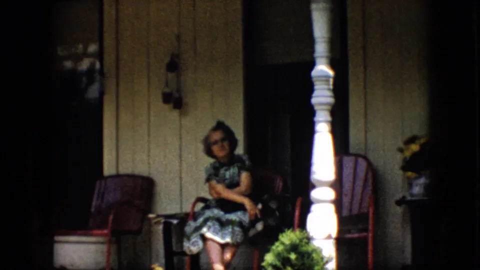 A woman in a patterned dress and glasses smiles from a red porch chair on a shaded Pennsylvania front porch in 1958 home-movie footage.
