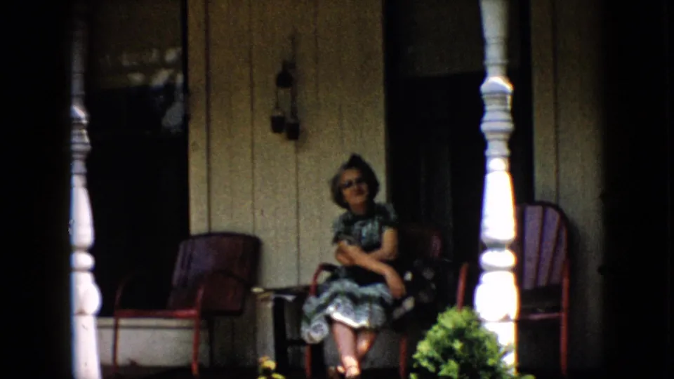 The frame shows a woman seated beside red porch chairs, white columns, wood siding, and a wall lantern in the 1958 Pennsylvania porch scene.
