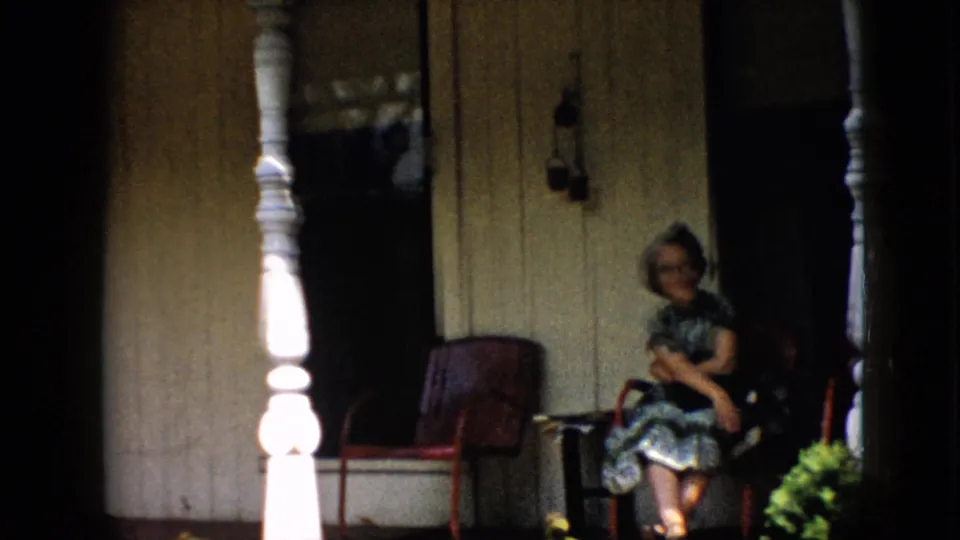 A woman sits near a white porch post with red chairs, wood siding, a wall lantern, and greenery visible in the 1958 Pennsylvania home-movie frame.