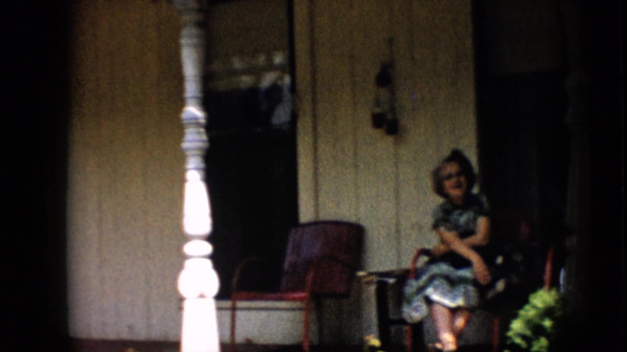 1958 Pennsylvania home-movie frame showing a woman in a patterned dress seated on a porch, with a white post in the foreground and a shadowed doorway behind her.