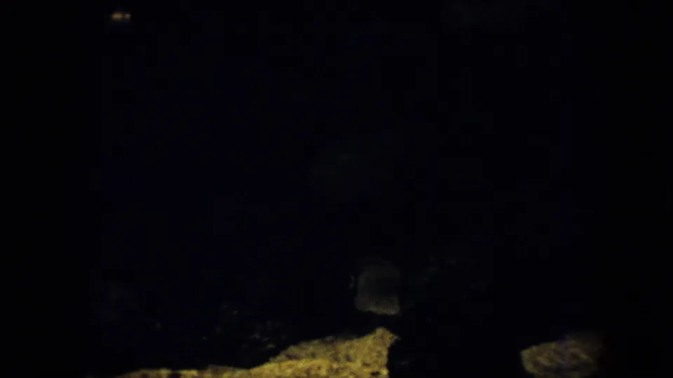 Very dark 1958 Pennsylvania 8mm frame showing a small fish-like shape near a pale rock area, with most of the view lost in underwater shadow.