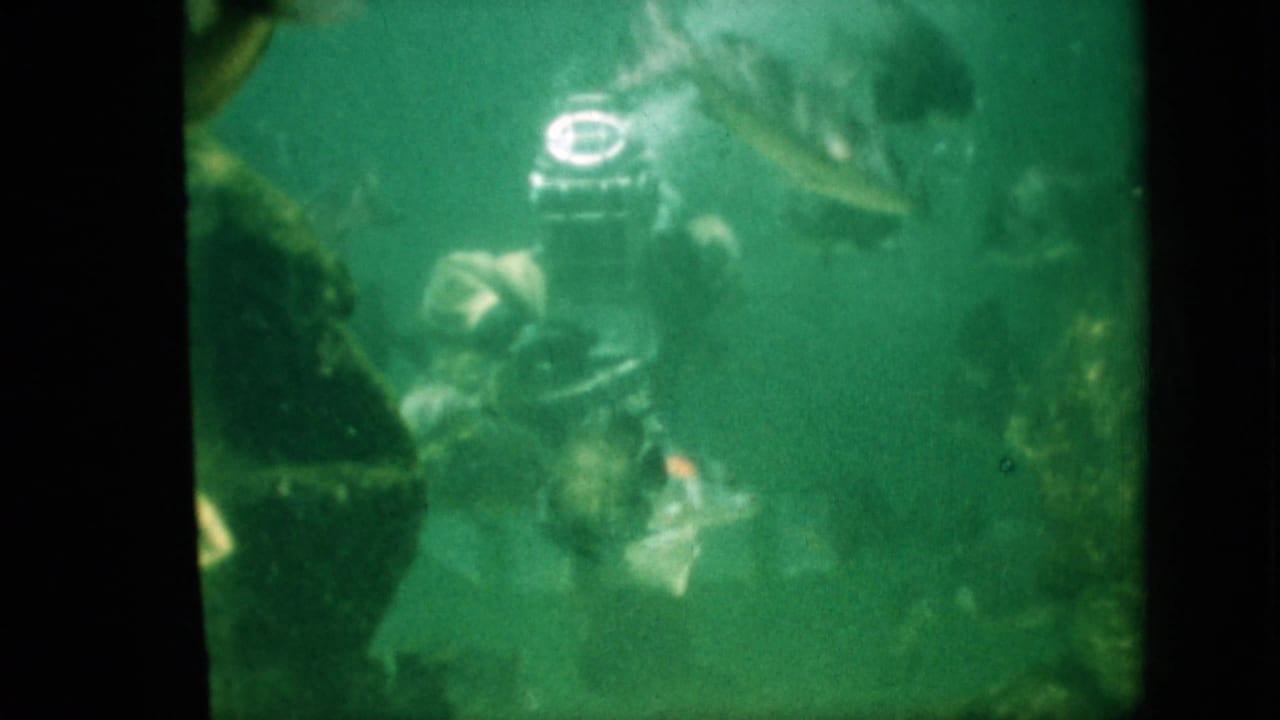 1958 Pennsylvania aquarium footage frame with a helmeted diver in a heavy suit moving near rocks as fish swim through green water.