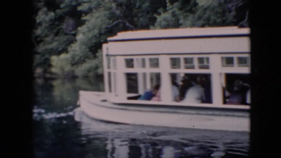 A white enclosed tour boat passes along a calm tree-lined river in Pennsylvania, with seated passengers visible through the cabin windows in 1958 home-movie footage.