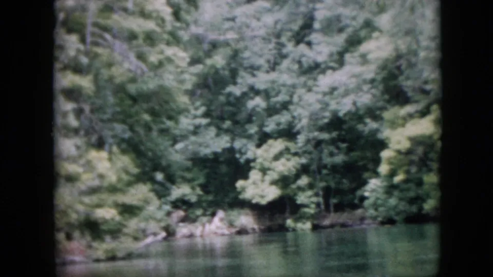 1958 Pennsylvania 8mm frame with green river water in the foreground and leafy woodland, shoreline rocks, and shaded vegetation beyond.