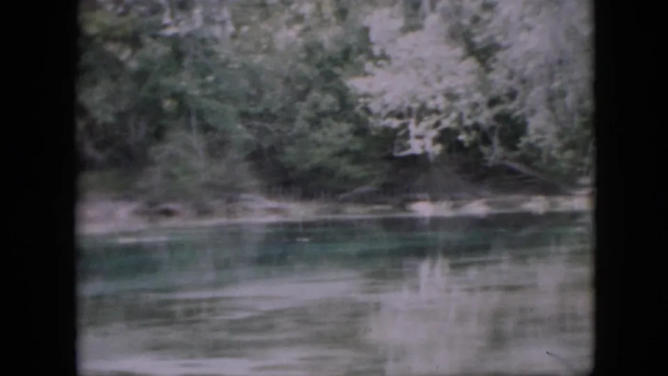 1958 Pennsylvania 8mm frame showing calm green-blue water, faint reflections, and a dense wooded shoreline viewed from the water.