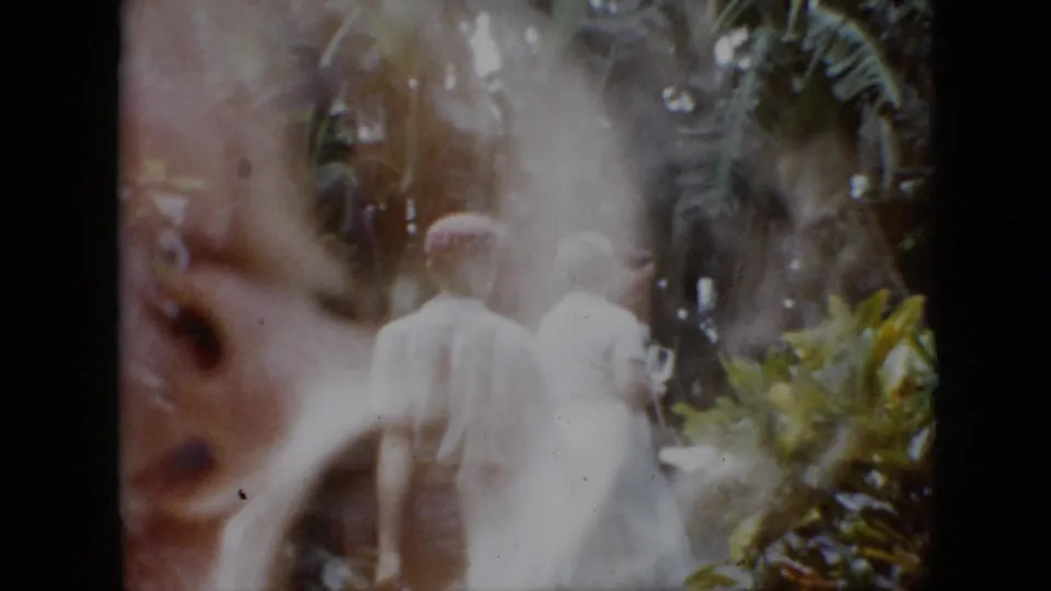 In 1958 Pennsylvania home-movie footage, an adult man in a red cap and an adult woman in a white dress walk away through lush garden plantings under soft, reflected 8mm light.