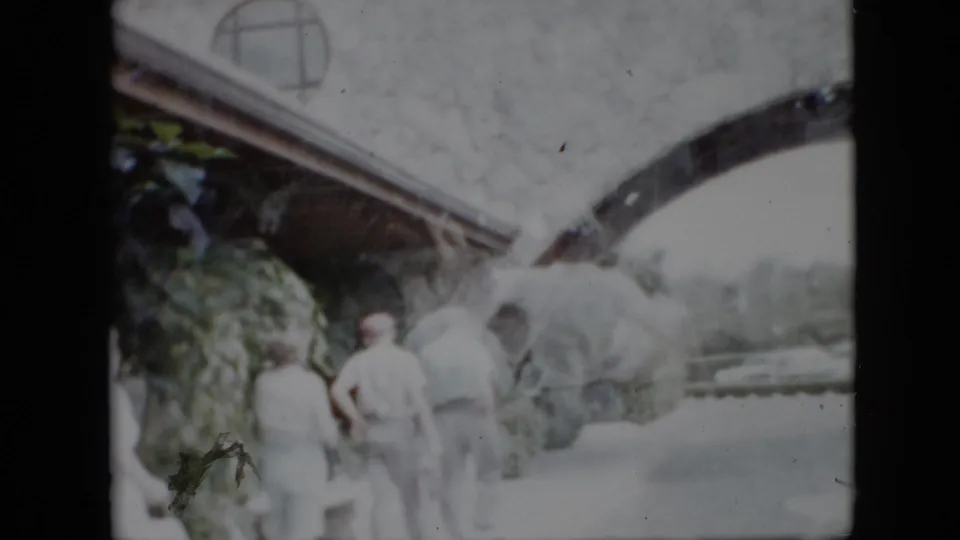 Several adults stand or walk near a pale stone archway or garden structure, with foliage at the edge of the soft, overexposed 8mm frame.
