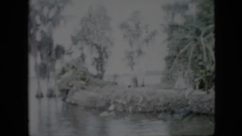 The frame shows a faded waterside landscape with calm water, trees, a planted bank, and distant figures in the supplied 1958 Pennsylvania home-movie clip.