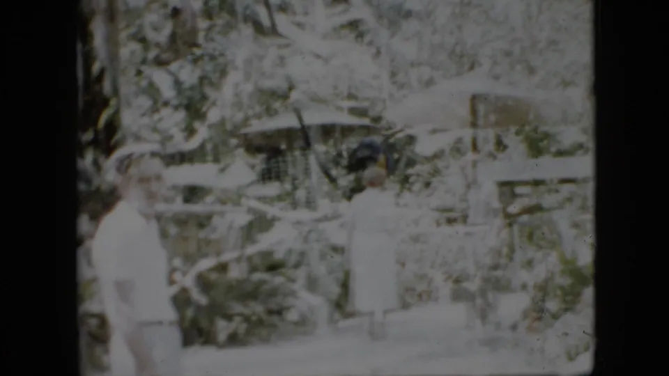 A 1958 Pennsylvania 8mm frame shows adults moving through a planted outdoor area, with pale clothing, possible umbrellas, and bright faded exposure.
